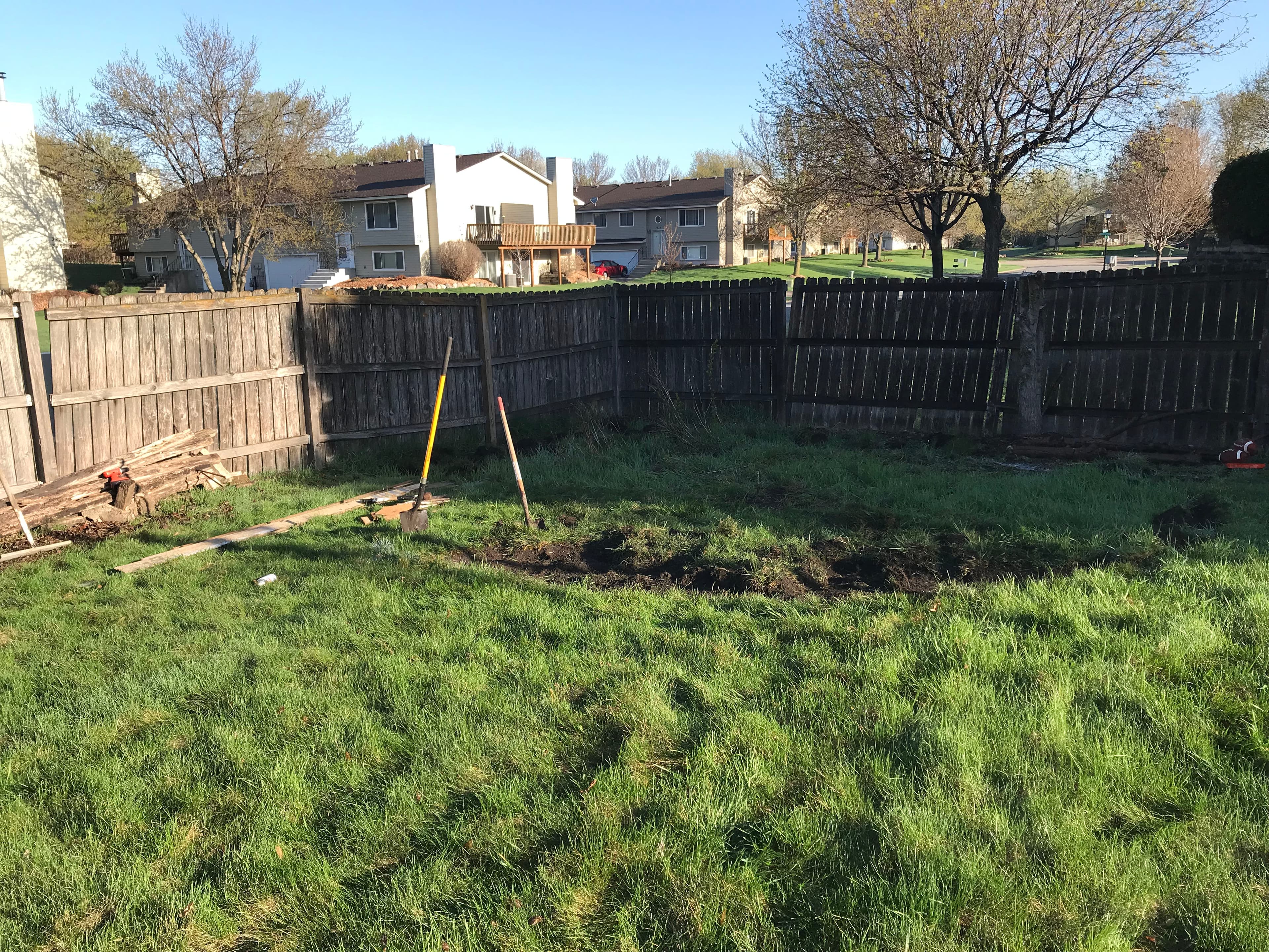 Backyard landscape project with a fenced area, tools, and freshly dug soil under a clear blue sky.