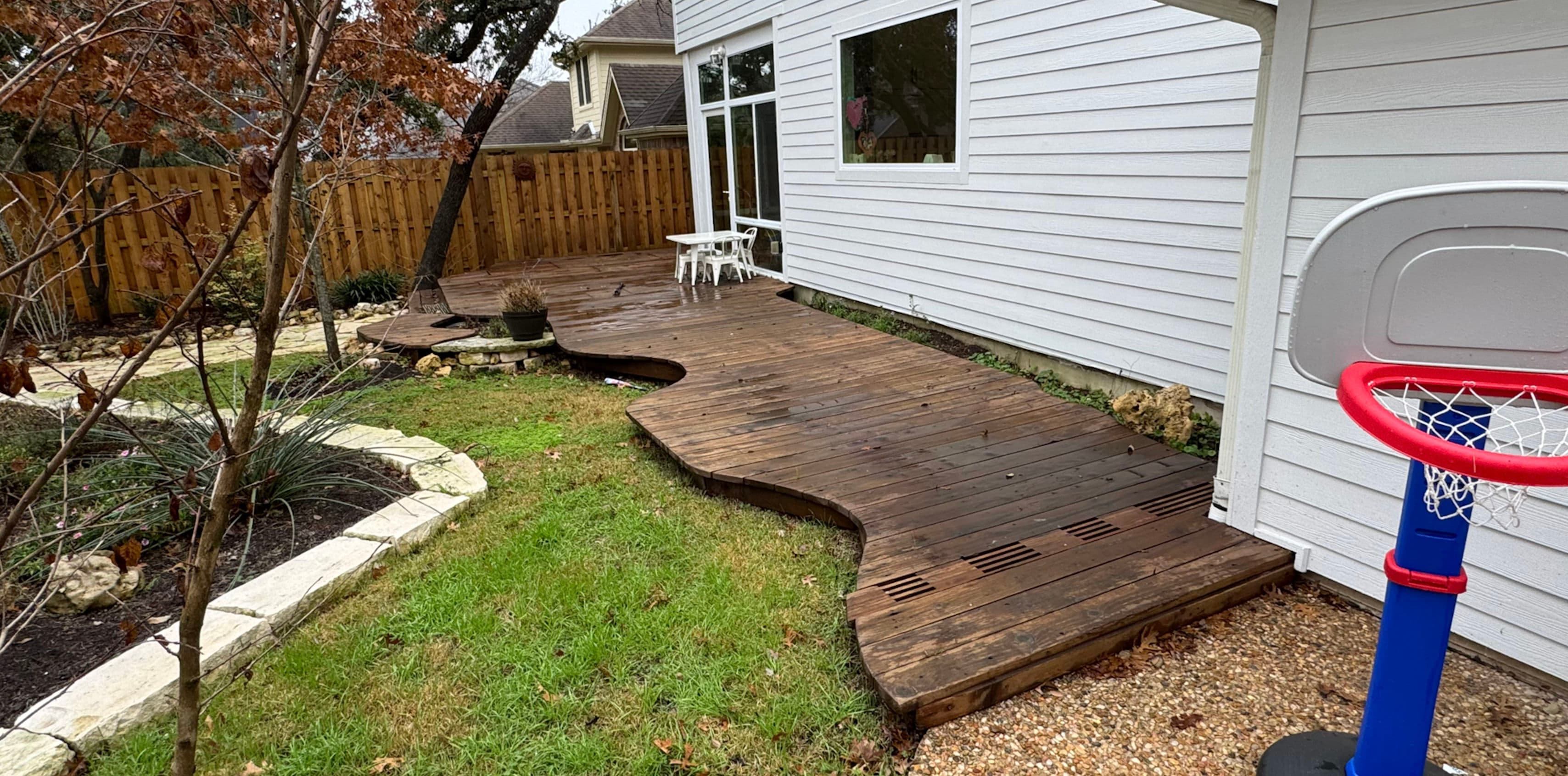 Curved wooden deck with green lawn, bordered by landscaping and a basketball hoop.