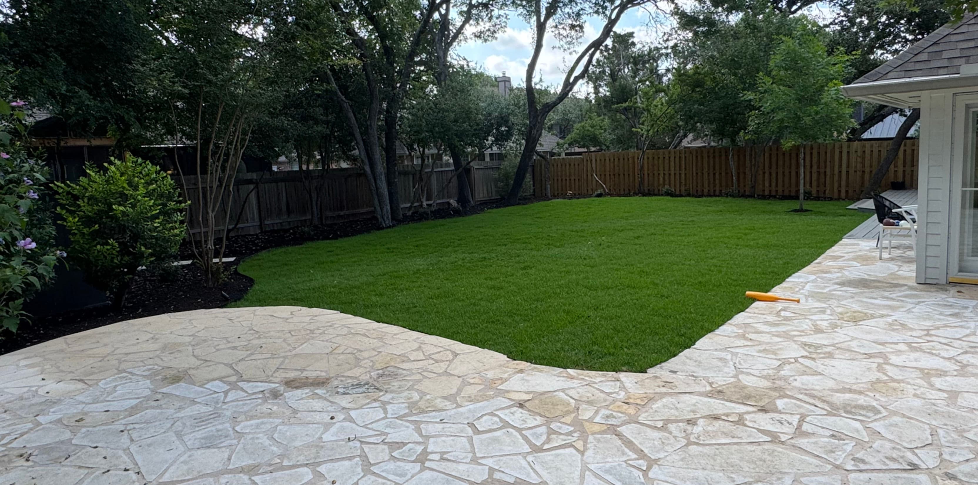 Lush green backyard with stone patio and wooden fence, surrounded by trees. Ideal outdoor space.