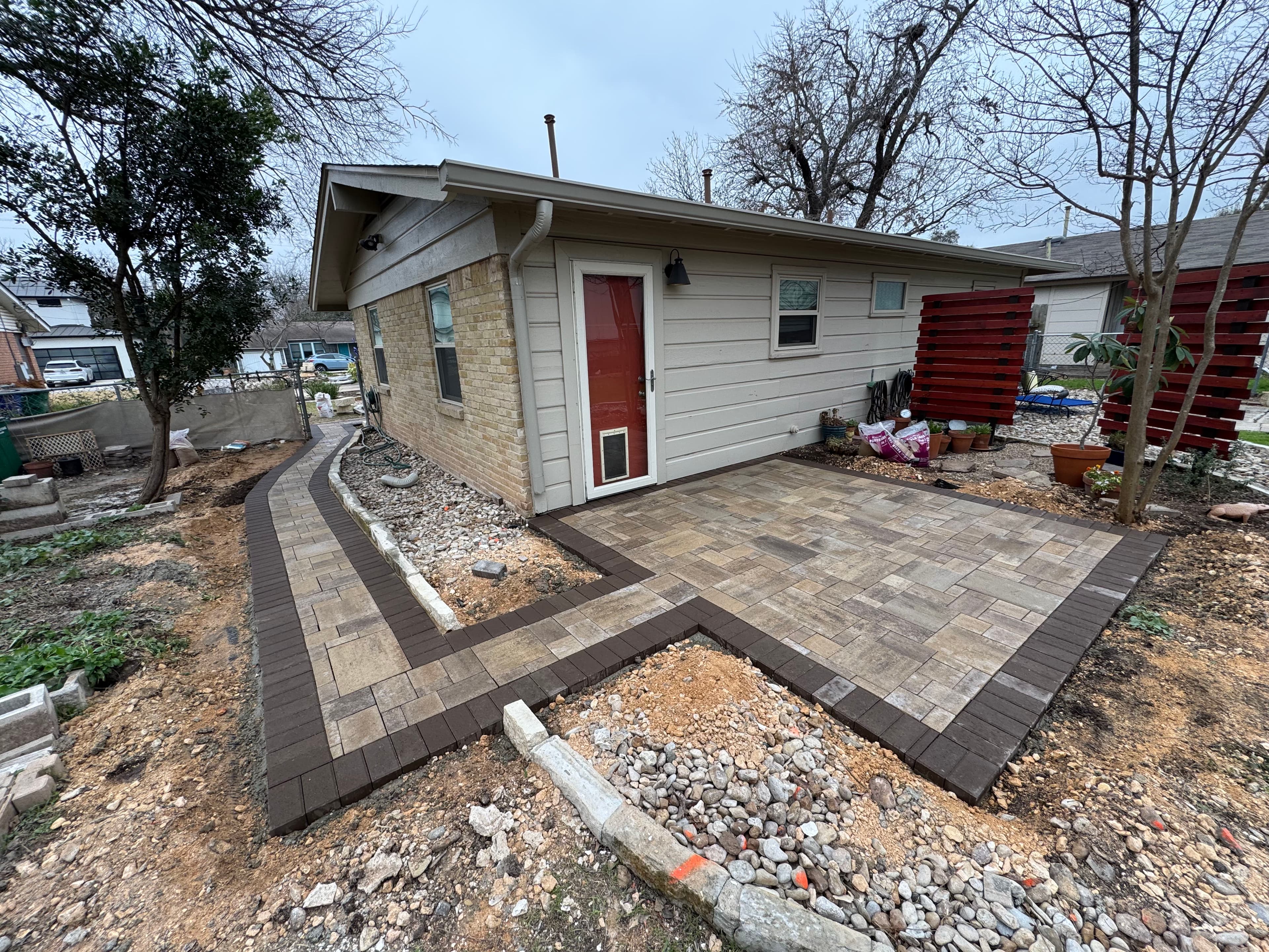 Paver patio and pathway leading to a small residential house in a landscaped yard.