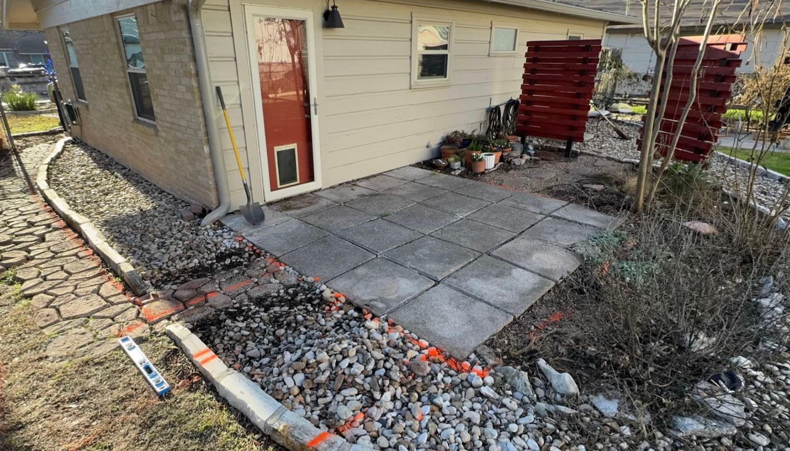 Paved patio area beside a house with garden beds and stones, ready for landscaping.