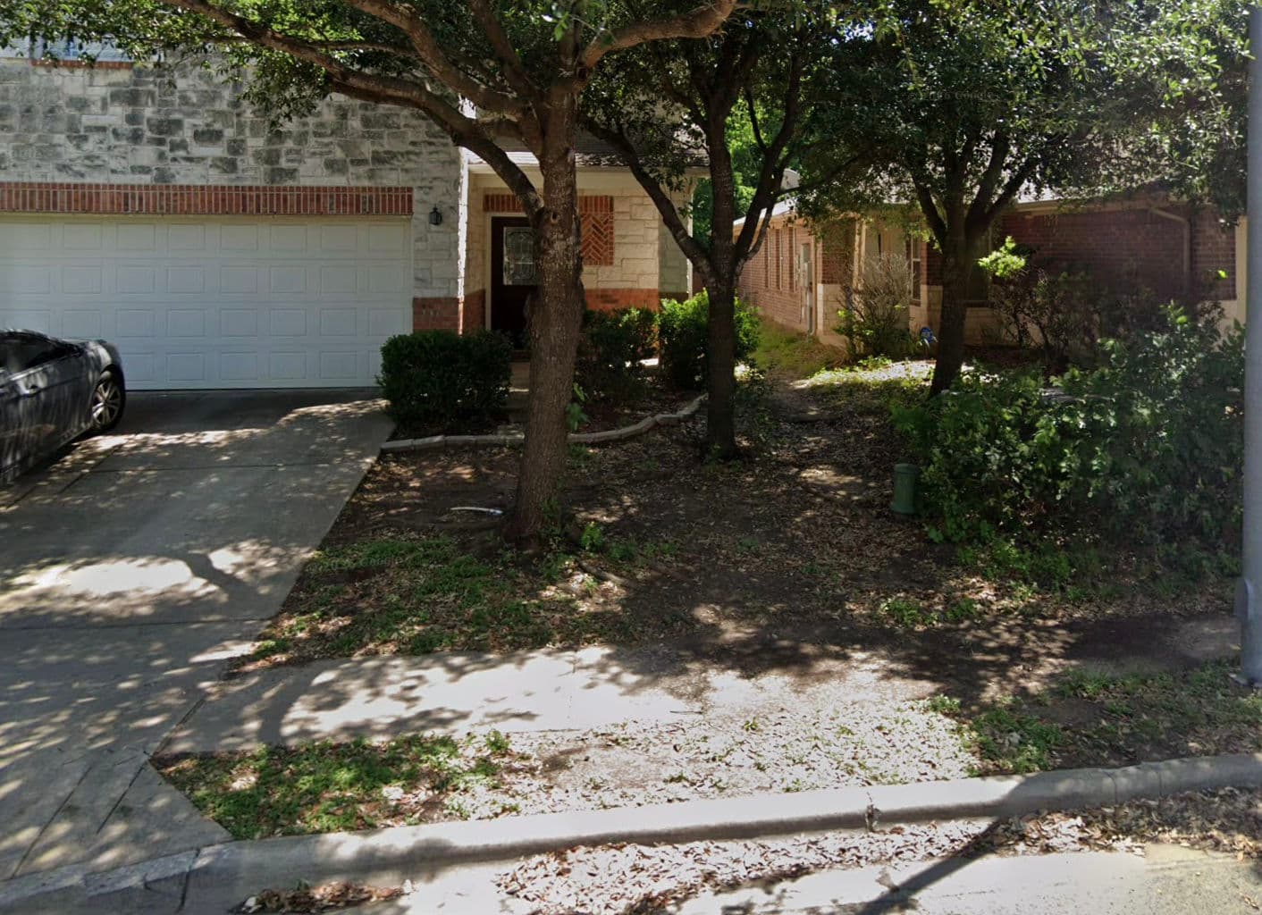 Residential landscape featuring a driveway, garage, and shaded front yard with trees.