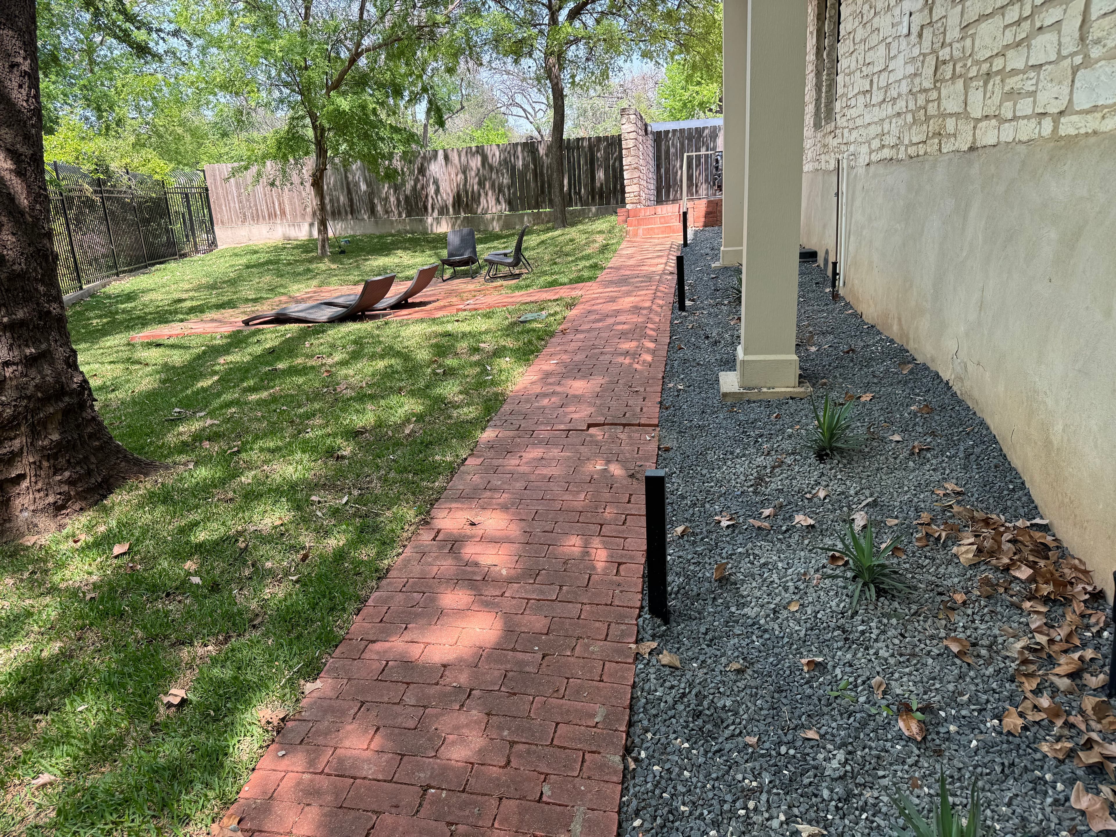 Brick pathway leading through a landscaped yard with gravel and plants. Sunny outdoor setting.