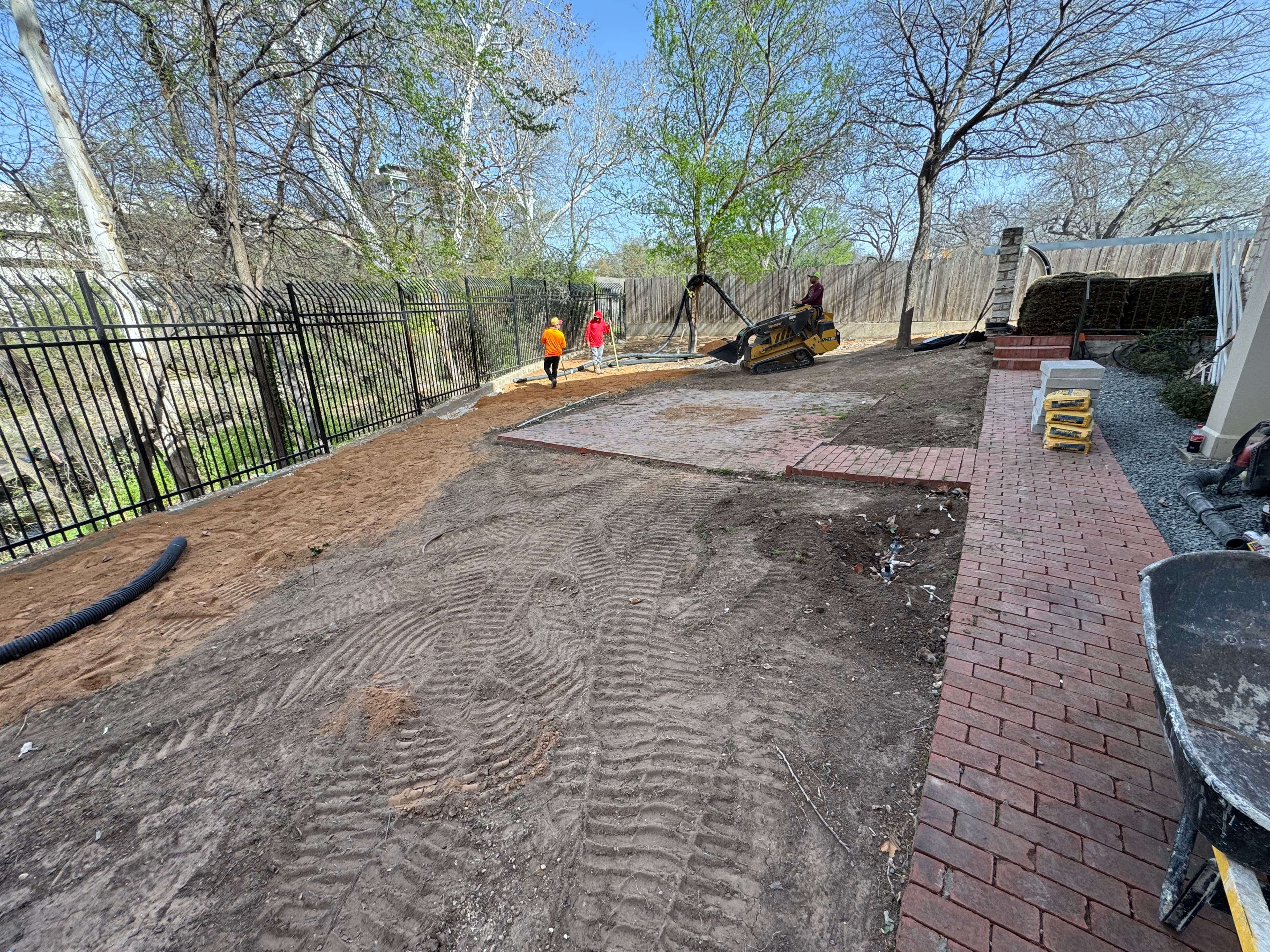 Construction workers grading a yard with machinery, surrounded by trees and a fence.