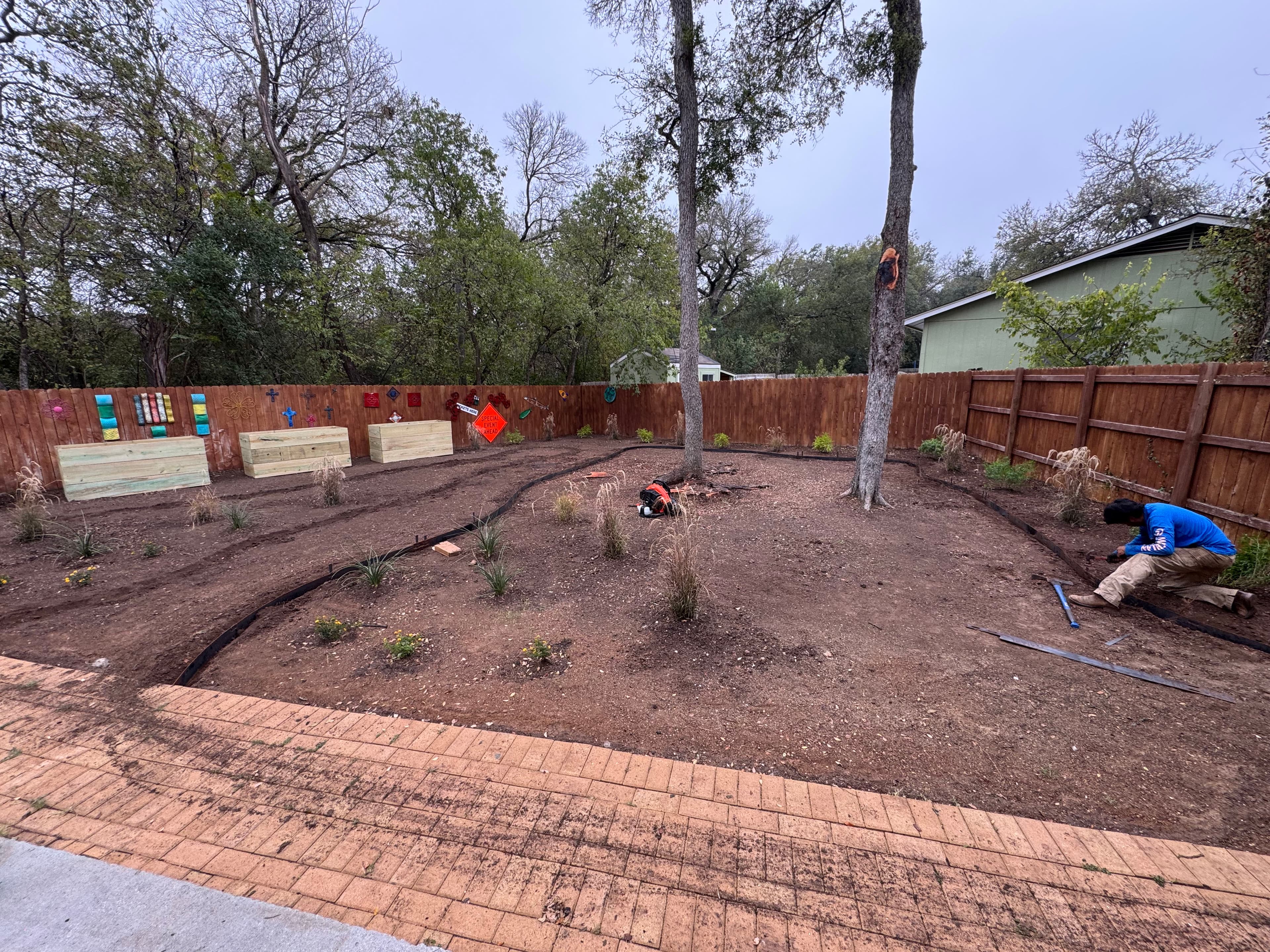 Person working in a landscaped garden with trees, flower beds, and a pathway.