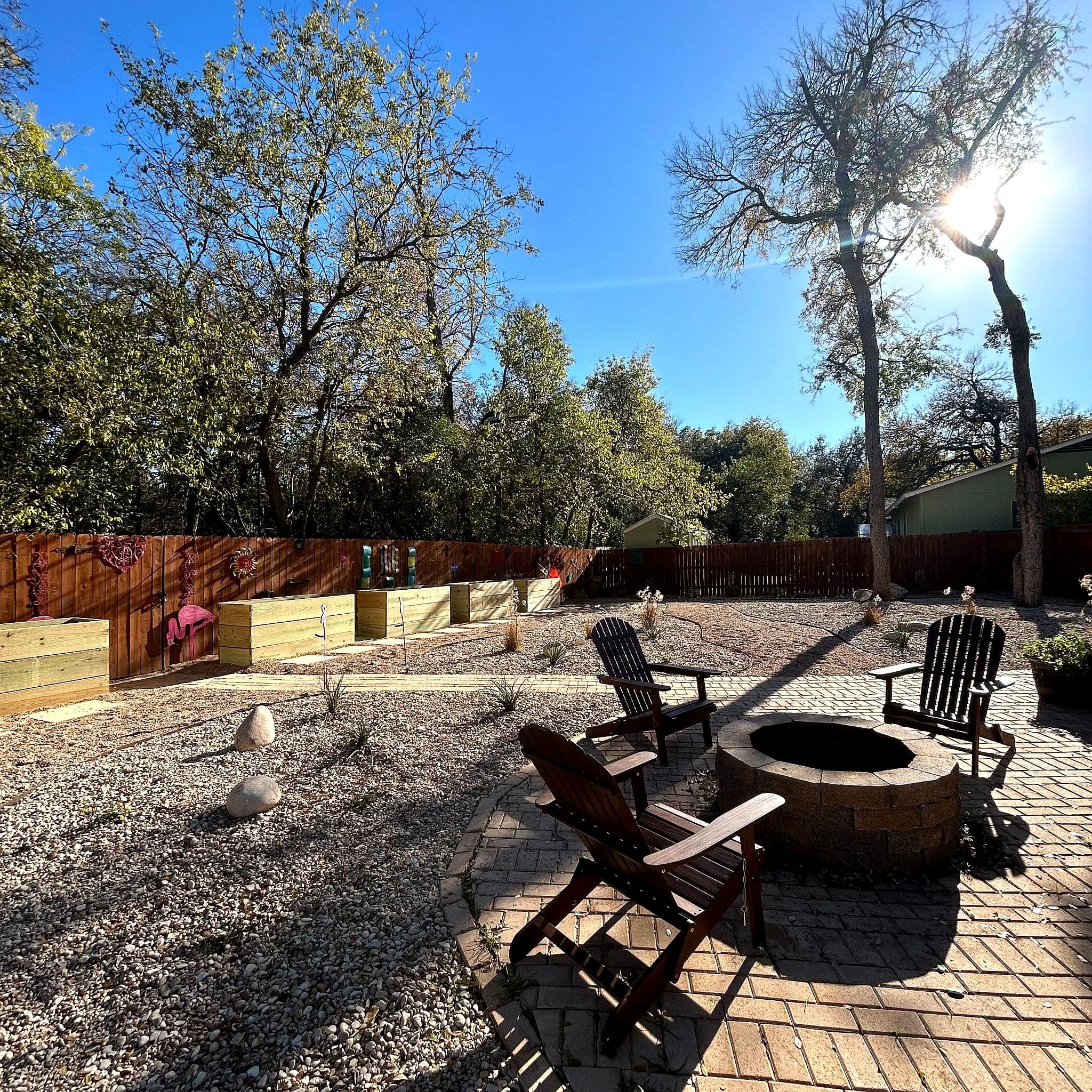 Outdoor seating area with fire pit, surrounded by trees and garden beds under a clear blue sky.