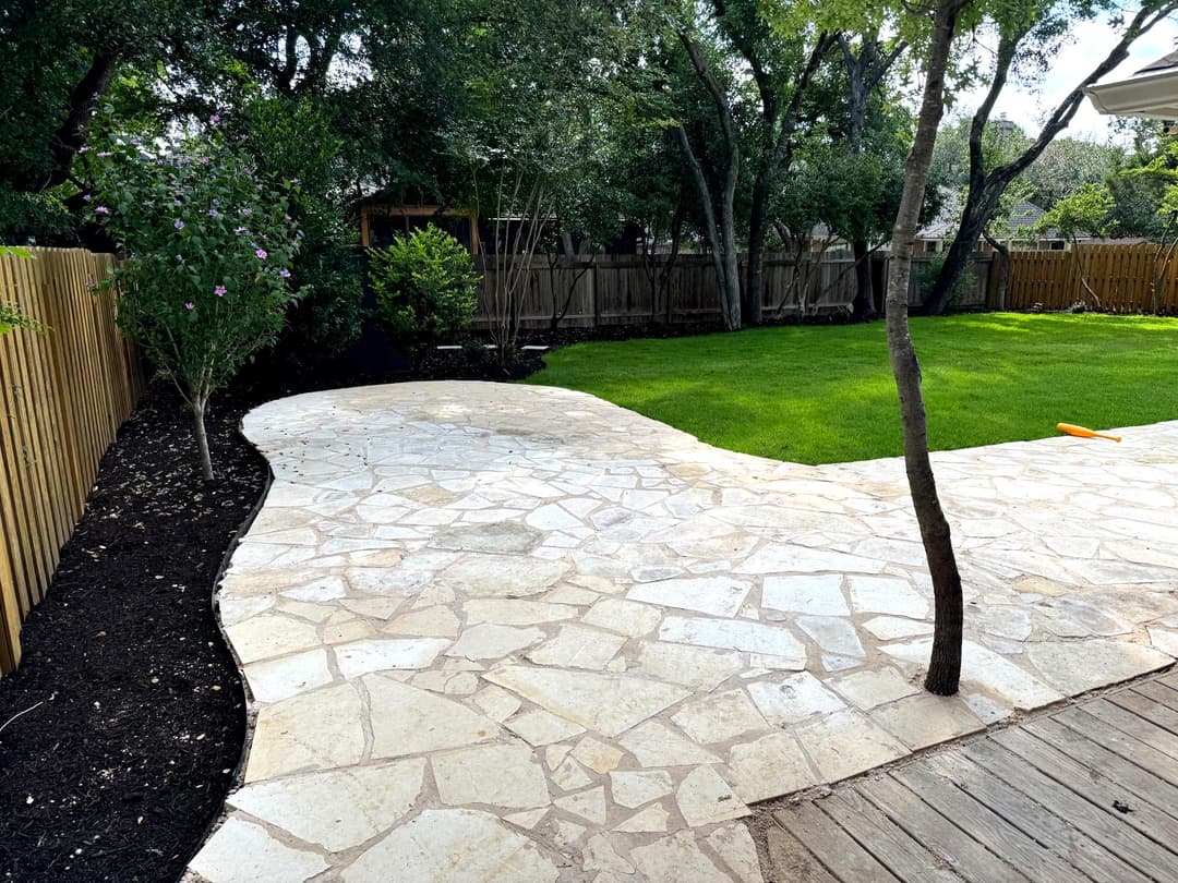 Stone pathway winding through a green backyard with trees and wooden fence.