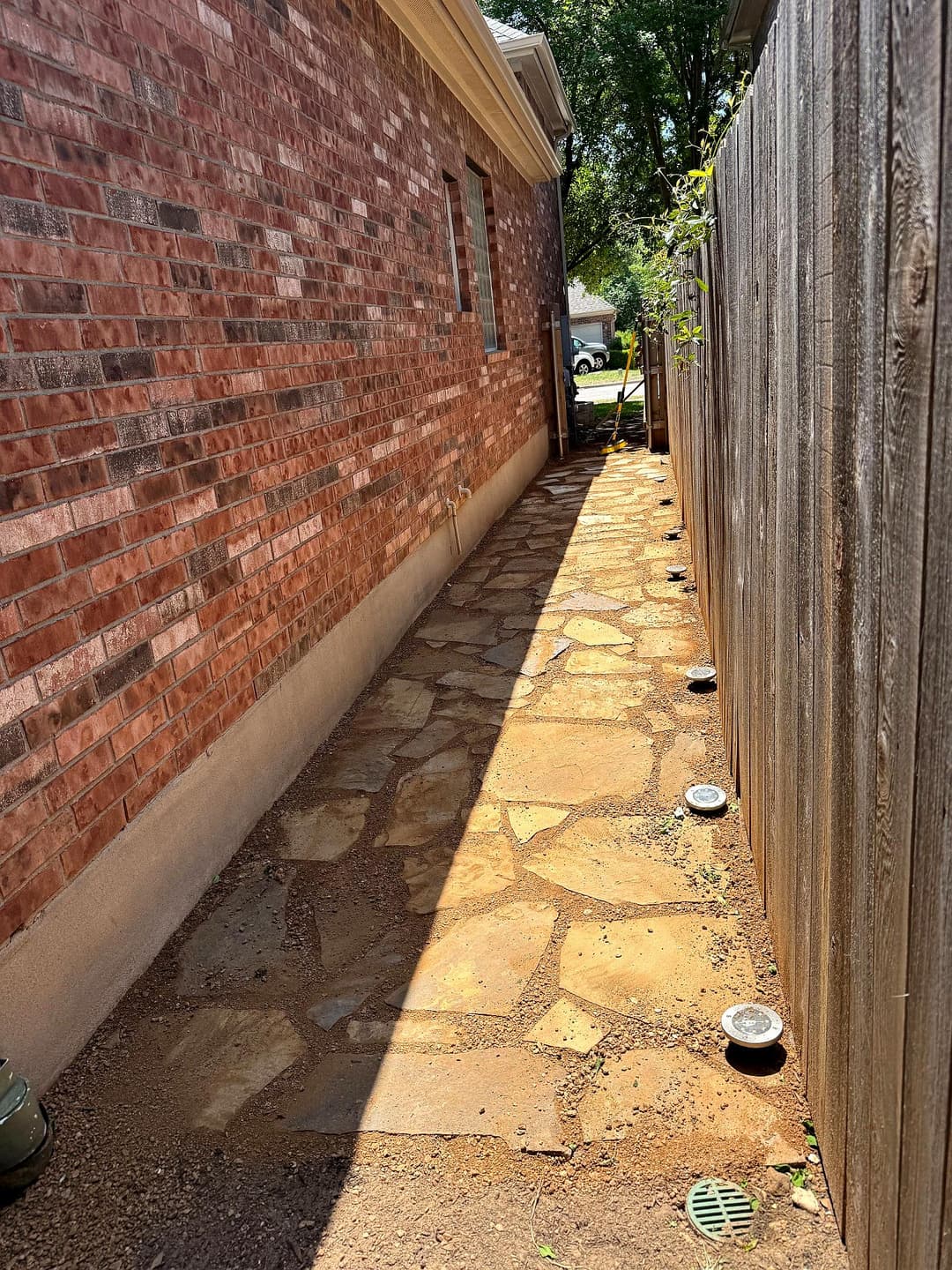 Narrow stone pathway beside a brick house, lined with ground lights and a wooden fence.