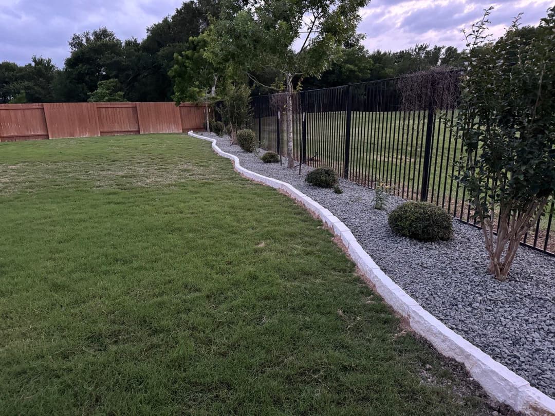 Lawn with landscaped gravel border and black fence in the background under a cloudy sky.