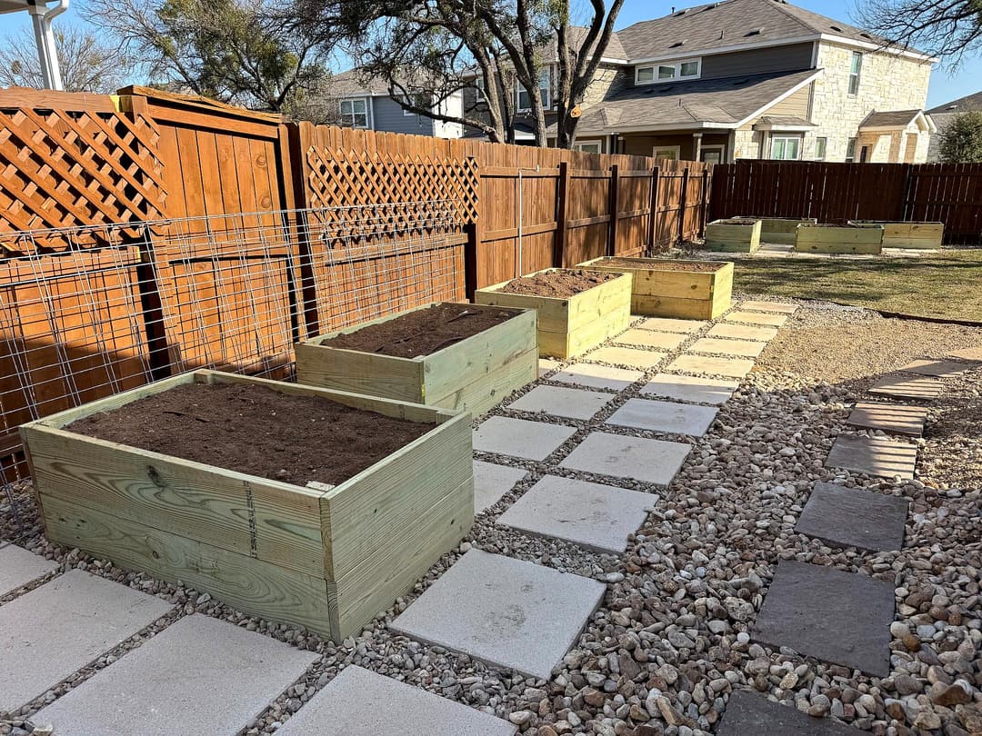 Wooden raised garden beds set in a backyard with stone pathways and fence in background.