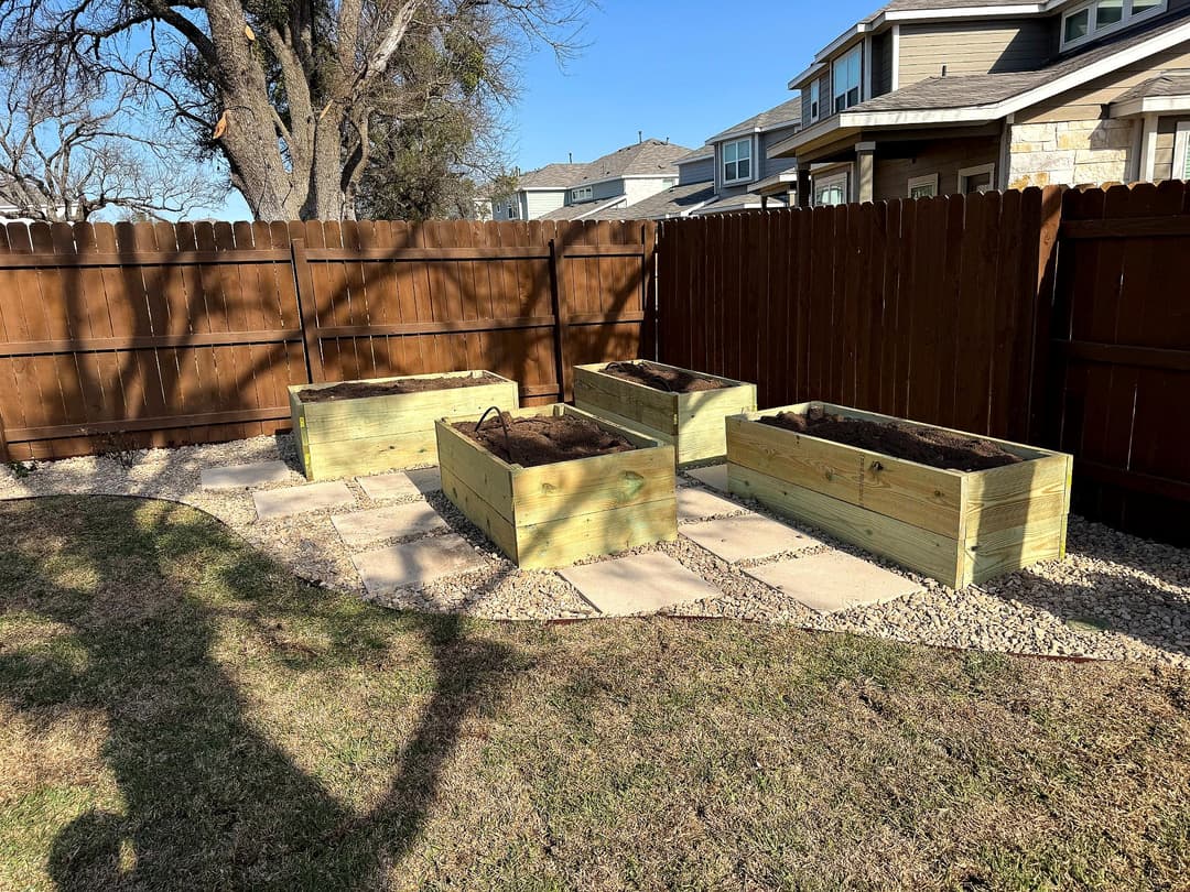 Wooden raised garden beds in a backyard with a fenced area and stone pathway.
