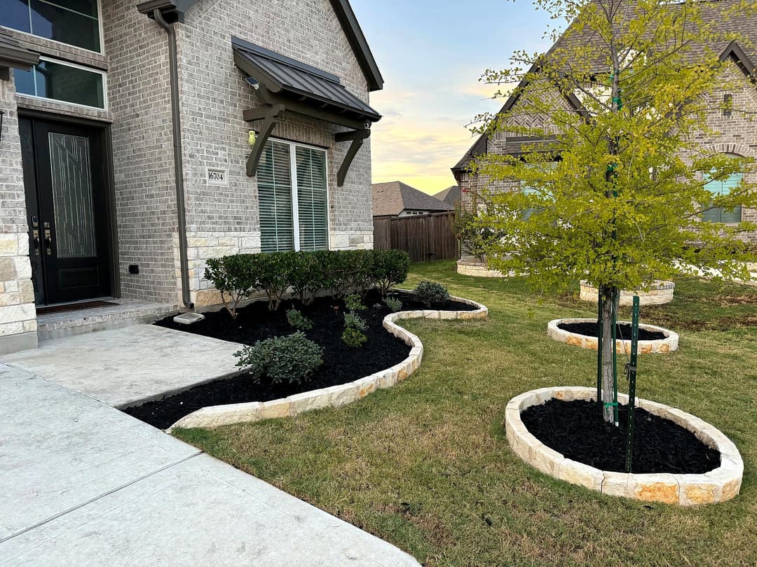 Landscaped front yard with stone-edged flower beds, shrubs, and a young tree.