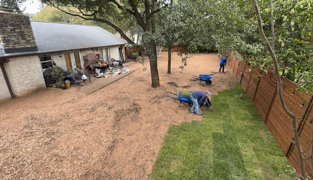 Workers installing sod in a backyard landscape with a stone house and wooden fence.