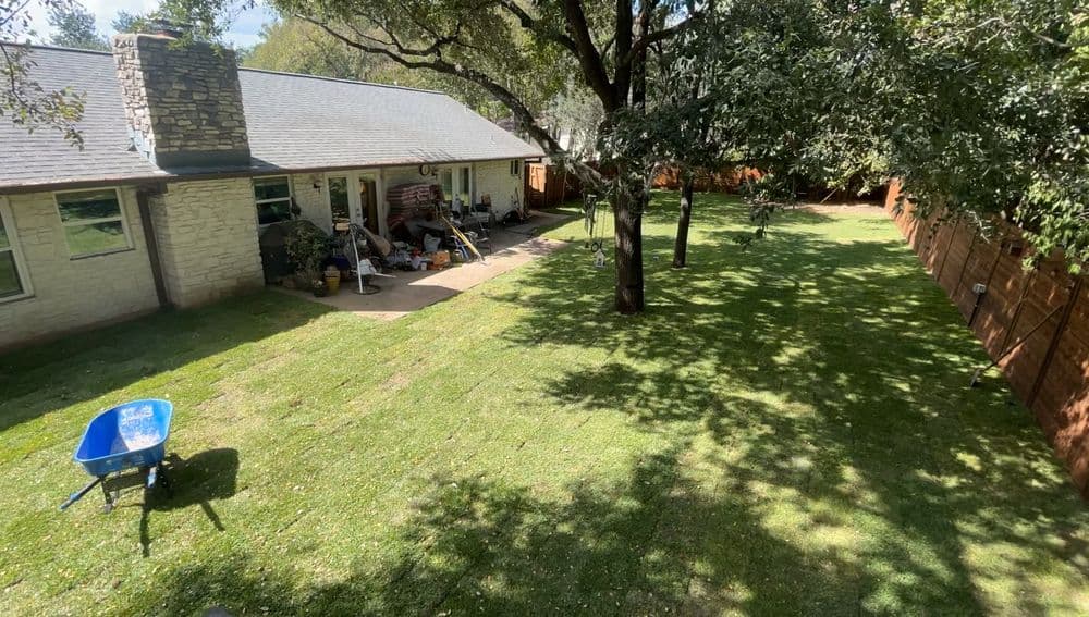 A spacious backyard with a well-maintained lawn, trees, and a wheelbarrow near a house.